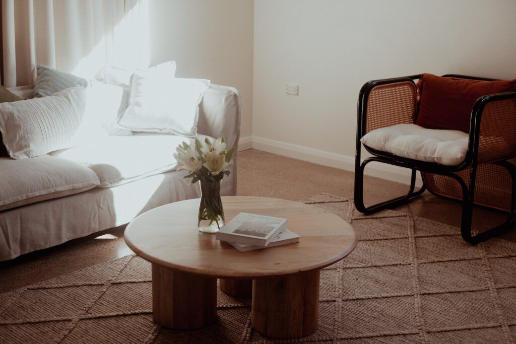 Living room with couch, pillows, and curtains, with circular table in middle with vase of flowers near chair