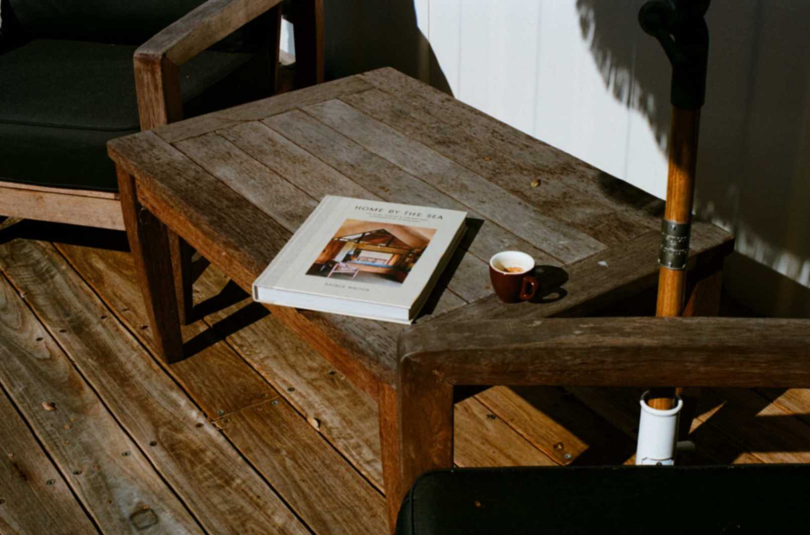 Home By The Sea Book on table near brown coffee mug and outdoor chairs under umbrella